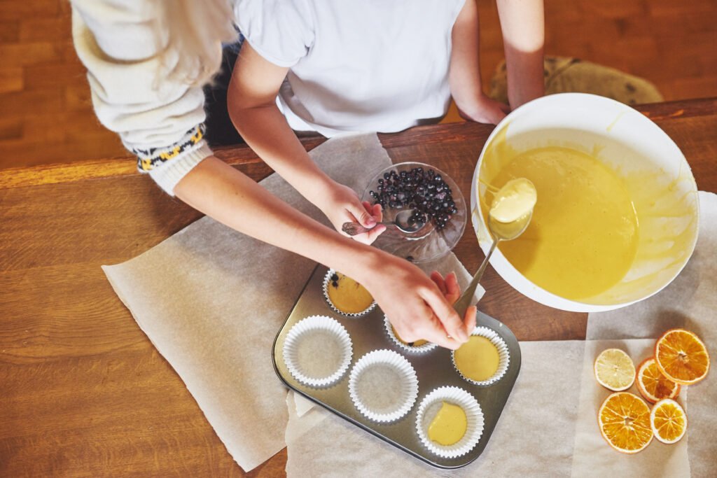 Children preparing pumpkin muffin recipe two ingredients by spooning batter into cupcake liners, viewed from above.